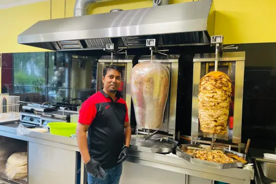 Mężczyzna w kuchni z dwoma obrotowymi rożnami z mięsem. Obok niego stos placków do kebaba. To właśnie kraftowe mięso kebab, co to znaczy? To świeżość i smak!